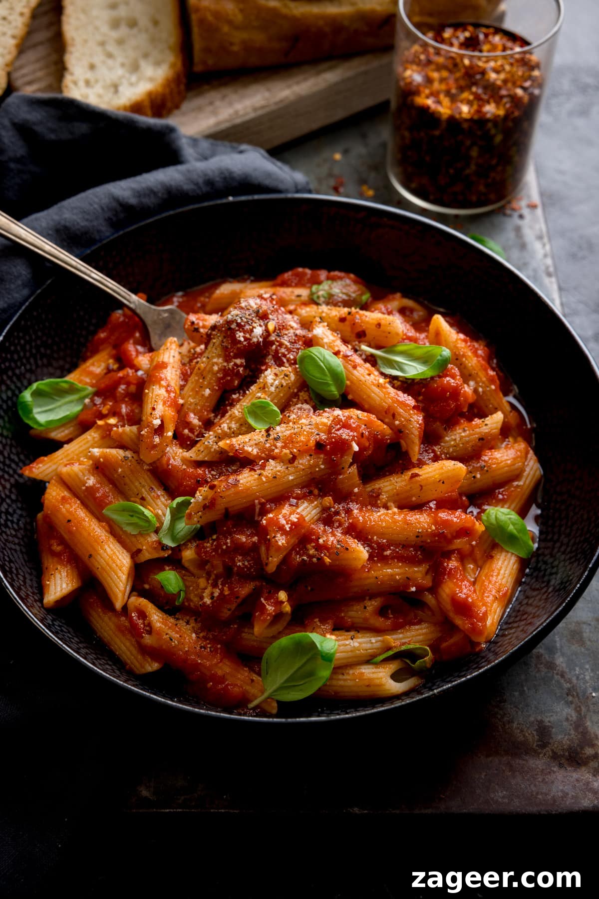 Spicy Penne Arrabbiata Recipe with Garlic and Tomato Sauce 2 A black bowl filled with penne arrabiata, with fresh baby basil leaves sprinkled on top. There is a fork sticking out of the bowl. The bowl is in a dark surface and there is a loaf of bread with a couple of sliced pieces and a jar of chilli flakes at the top of the frame.