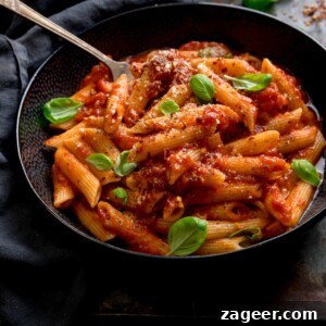 Spicy Penne Arrabbiata Recipe with Garlic and Tomato Sauce 13 A black bowl filled with penne arrabiata, with fresh baby basil leaves sprinkled on top. There is a fork sticking out of the bowl. The bowl is in a dark surface, next to a grey napkin.