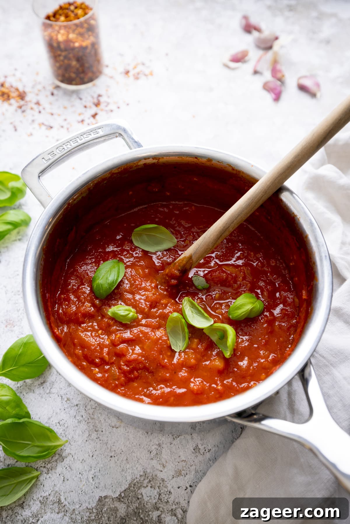 Spicy Penne Arrabbiata Recipe with Garlic and Tomato Sauce 4 Overhead image of a silver pan filled with arrabbiata sauce on a light background. There is a wooden spoon and sprinkled fresh basil leaves in the sauce. There are basil leaves, garlic cloves and a jar of chilli flakes around the pan.