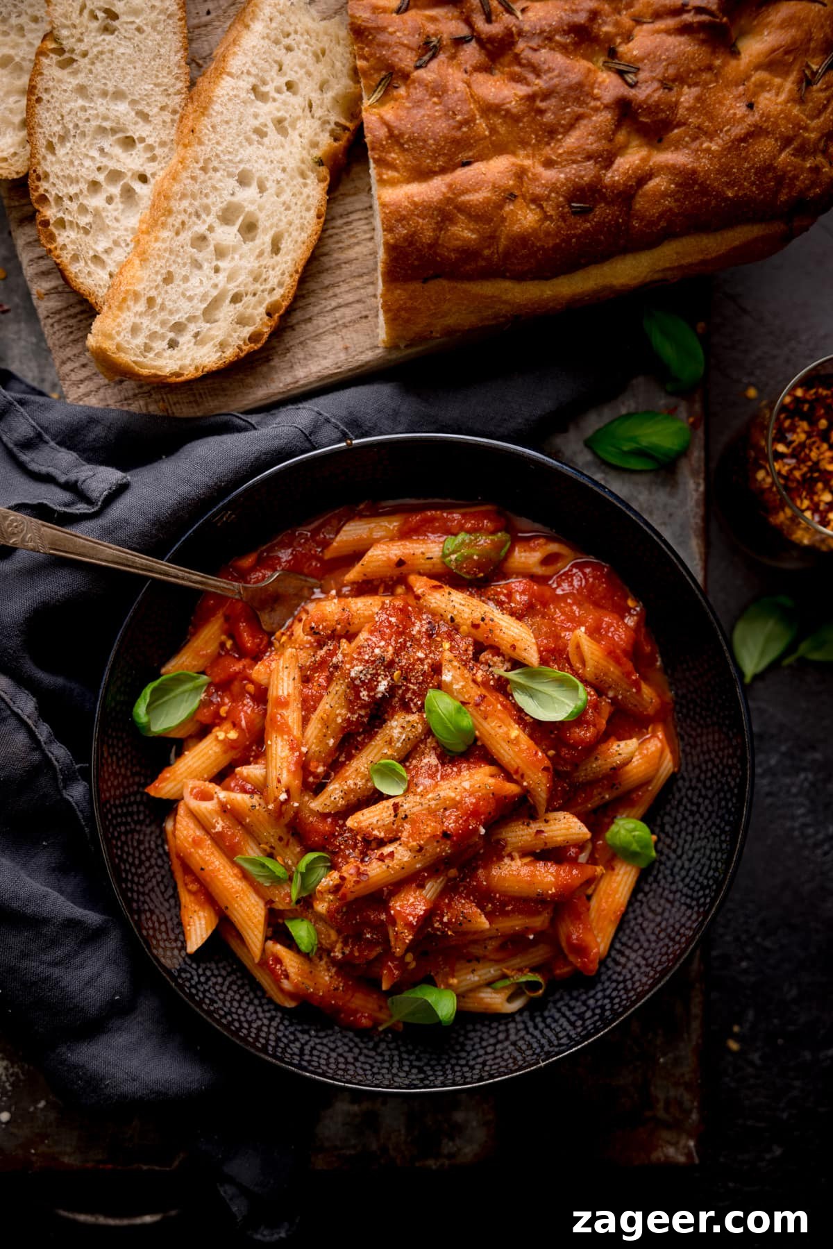 Spicy Penne Arrabbiata Recipe with Garlic and Tomato Sauce 5 Overhead image of a black bowl filled with penne arrabiata, with fresh baby basil leaves sprinkled on top. There is a fork sticking out of the bowl. The bowl is in a dark surface and there is a loaf of bread with a couple of sliced pieces at the top of the frame.