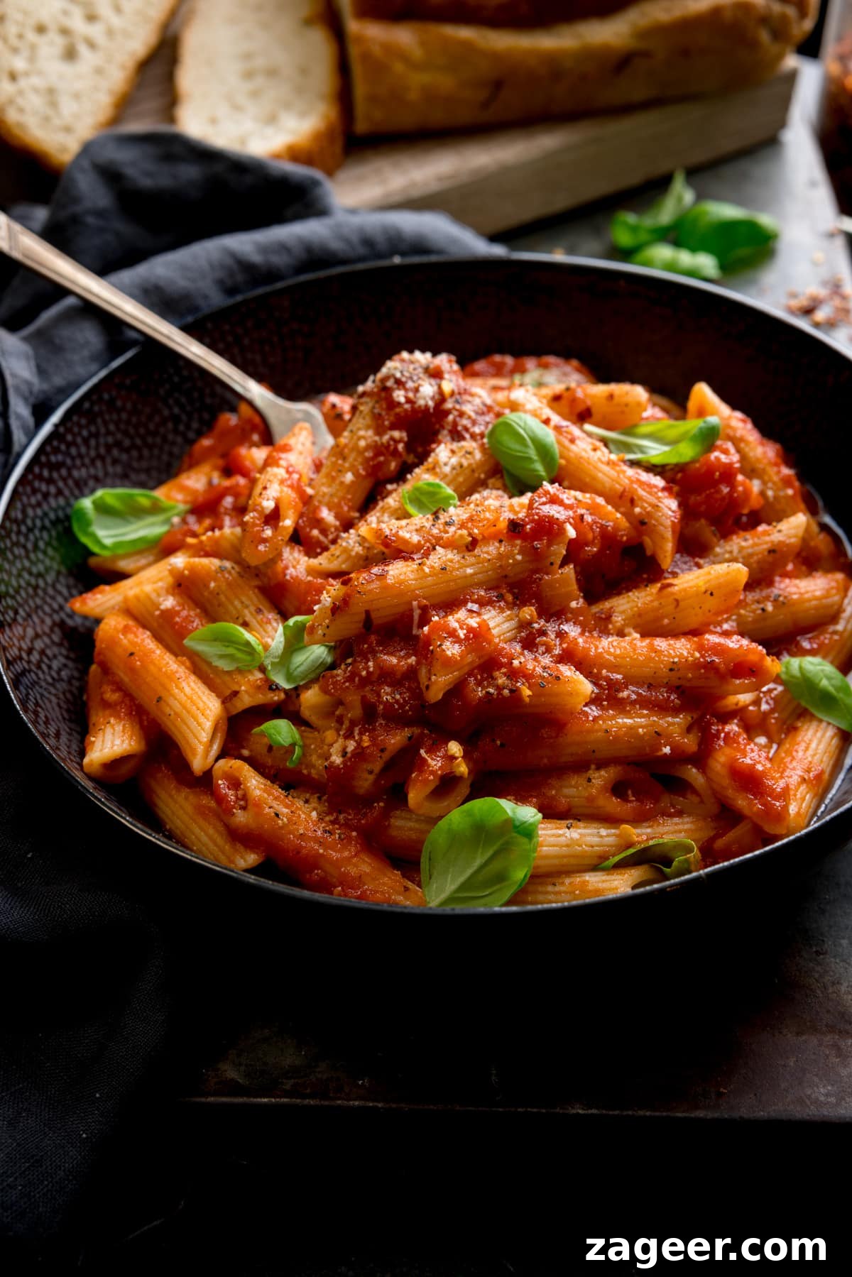 Spicy Penne Arrabbiata Recipe with Garlic and Tomato Sauce 6 A black bowl filled with penne arrabiata, with fresh baby basil leaves sprinkled on top. There is a fork sticking out of the bowl. The bowl is in a dark surface and there is a loaf of bread with a couple of sliced pieces at the top of the frame.