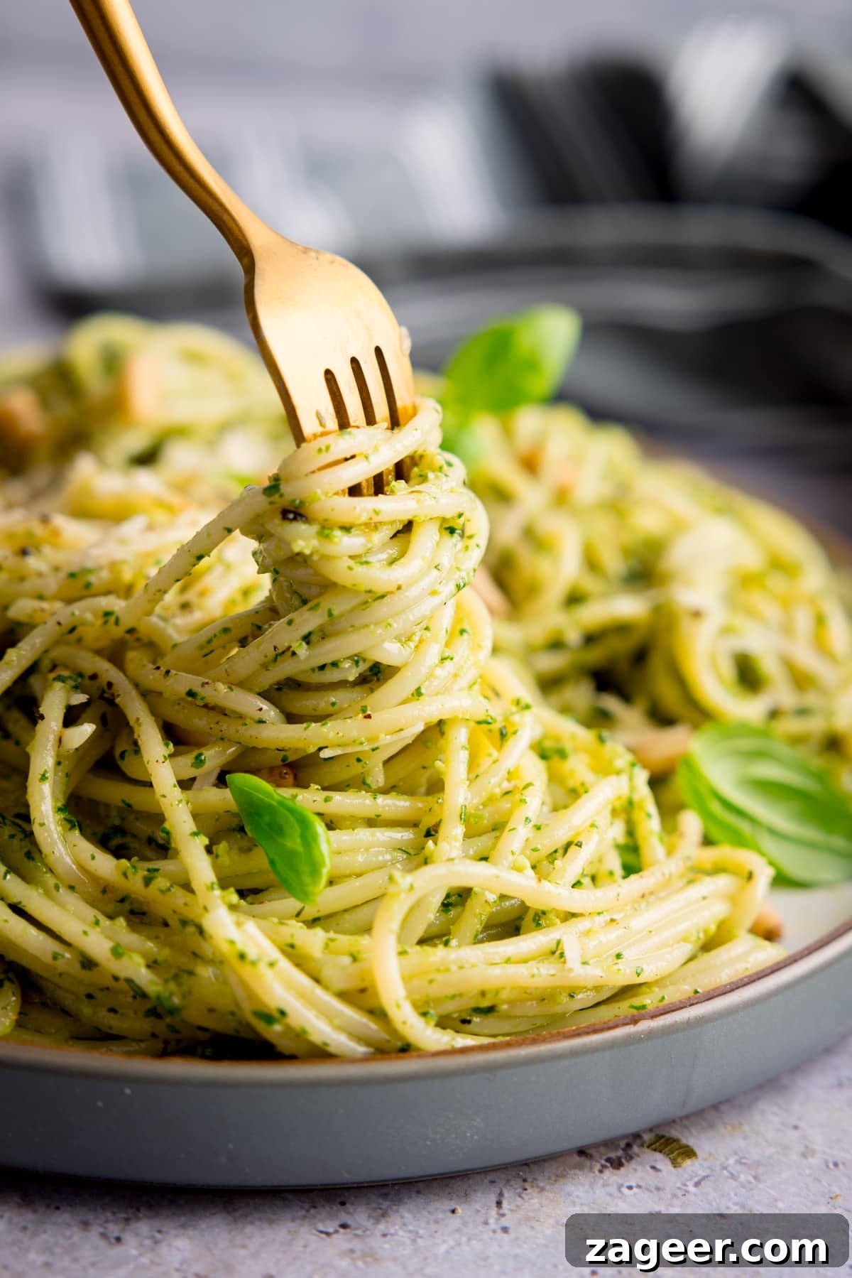 Creamy Basil Pesto Pasta with Garlic and Parmesan 2 Pesto spaghetti on a light grey plate being swirled with a gold fork. The pasta is sprinkled with baby basil leaves, toasted pine nuts and parmesan. The plate is on a light grey surface.
