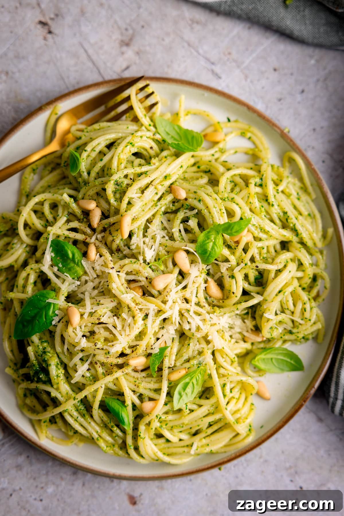 Creamy Basil Pesto Pasta with Garlic and Parmesan 5 Overhead image of pesto spaghetti on a light grey plate. The pasta is sprinkled with baby basil leaves, toasted pine nuts and parmesan. The plate is on a light grey surface and there is a gold fork on the plate.