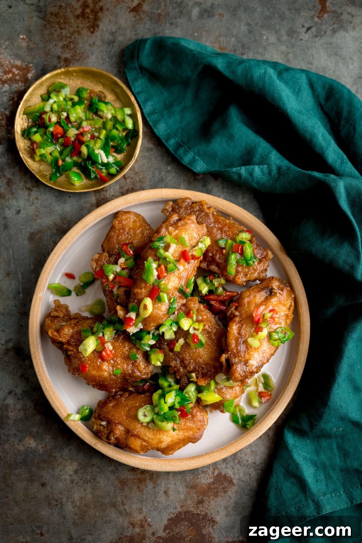 Crispy Salt and Pepper Chicken Wings Recipe 2 Overhead image of crispy salt and pepper chicken wings on a white plate on a rustic metal surface. There is a green napkin and a small gold dish with some of the chilli-salt-pepper topping in it.