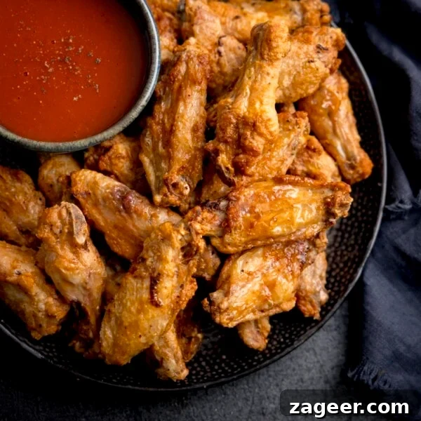 Crispy Salt and Pepper Chicken Wings Recipe 11 A square overhead shot of Air Fryer Crispy Chicken Wings piled up on a black speckled plate, their is also a large black bowl filled with buffalo sauce, sprinkled with seasoning on the plate, the plate is on a mottled dark grey background and there is some dark blue napkins to the side of the plate.