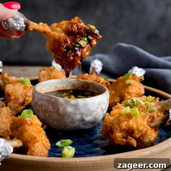 Crispy Salt and Pepper Chicken Wings Recipe 12 Square image of a plate of chicken lollipops with a bowl of spicy dipping sauce. One of the wings is being dipped into the sauce and is dripping. The wing is being held by fingers with pink-painted fingernails.