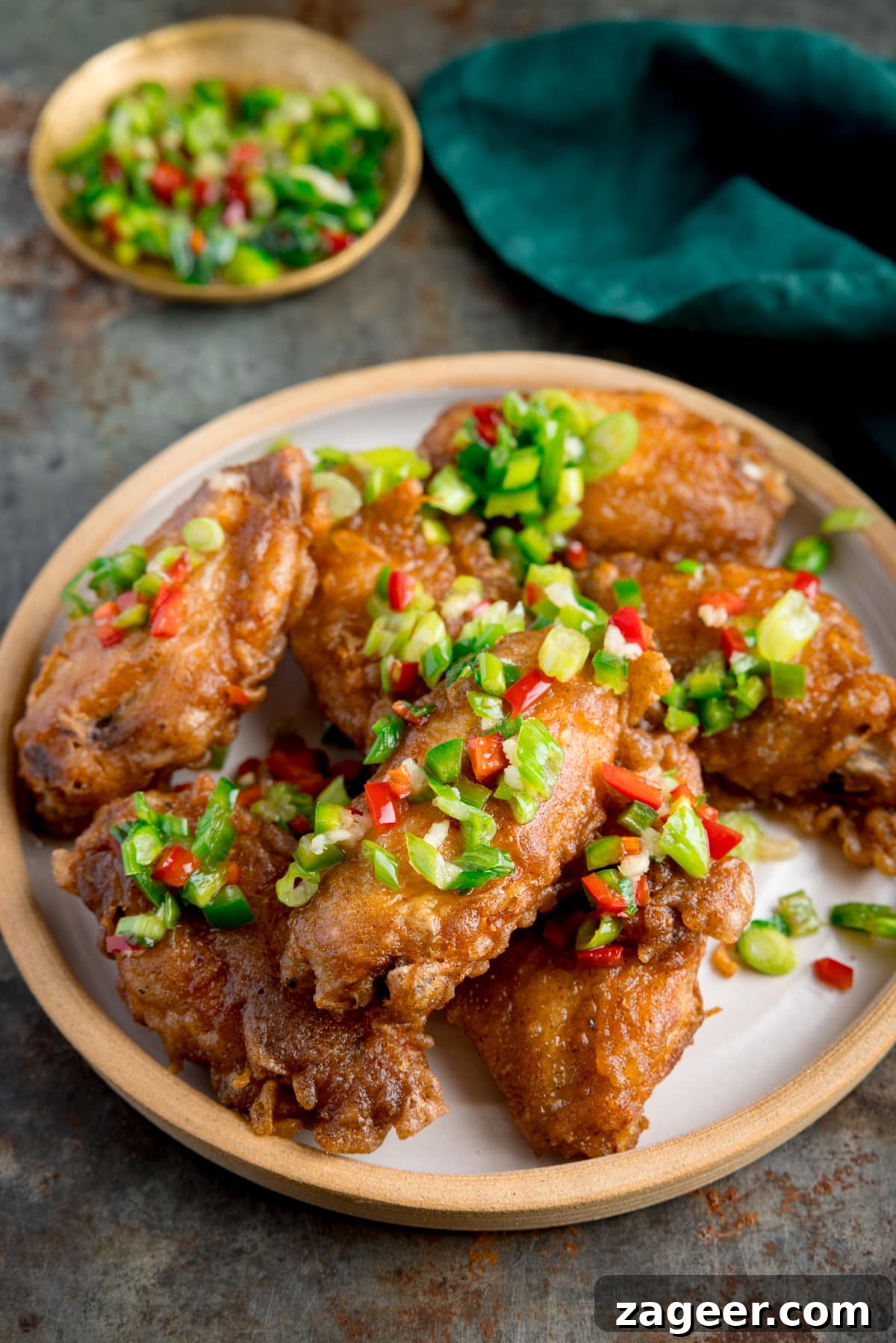 Crispy Salt and Pepper Chicken Wings Recipe 5 Tall side-on image of crispy salt and pepper chicken wings on a white plate on a rustic metal surface. There is a green napkin and a small gold dish with some of the chilli-salt-pepper topping in it at the top of the frame.
