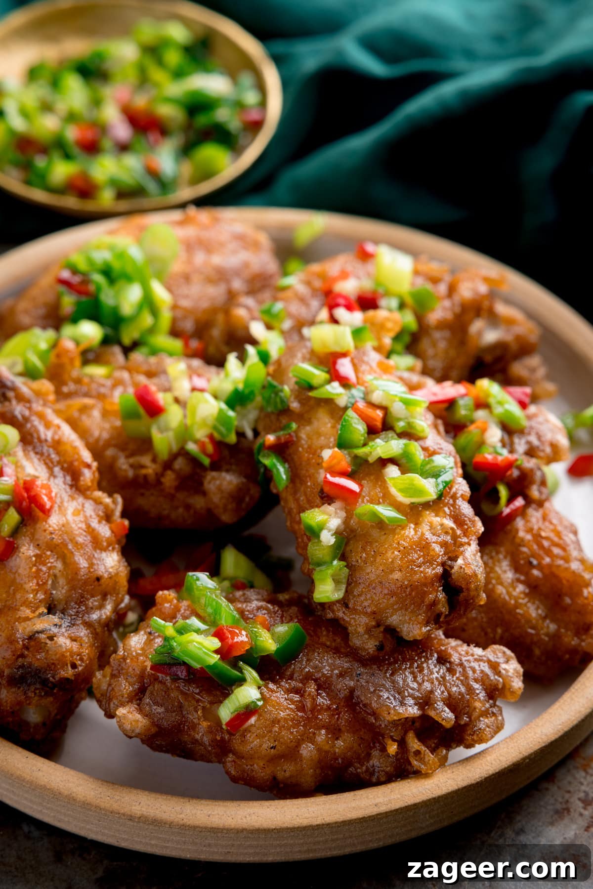 Crispy Salt and Pepper Chicken Wings Recipe 6 Tall side-on image of crispy salt and pepper chicken wings on a white plate on a rustic metal surface. There is a green napkin and a small gold dish with some of the chilli-salt-pepper topping in it at the top of the frame.
