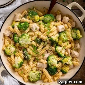 Overhead shot of chicken and broccoli pasta in a white cast iron pan with a wooden spoon sticking out. The pan is on a wooden table with a white napkin