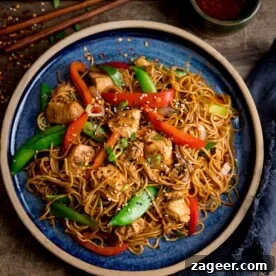 Overhead image of chicken lo mein on a blue plate on top of a wooden table. There is a pot of sweet chilli sauce and some chopsticks at the top of the image.