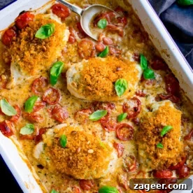 A white baking dish with four breaded chicken breasts in a creamy pesto sauce with roasted cherry tomatoes and basil leaves. There is a spoon resting in the top of the dish.