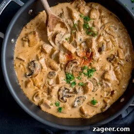 Square overhead image of chicken and mushroom stroganoff in a dark pan. The pan is on a dark surface with a wooden spoon sticking out. The stroganoff is garnished with chopped parsley and paprika.