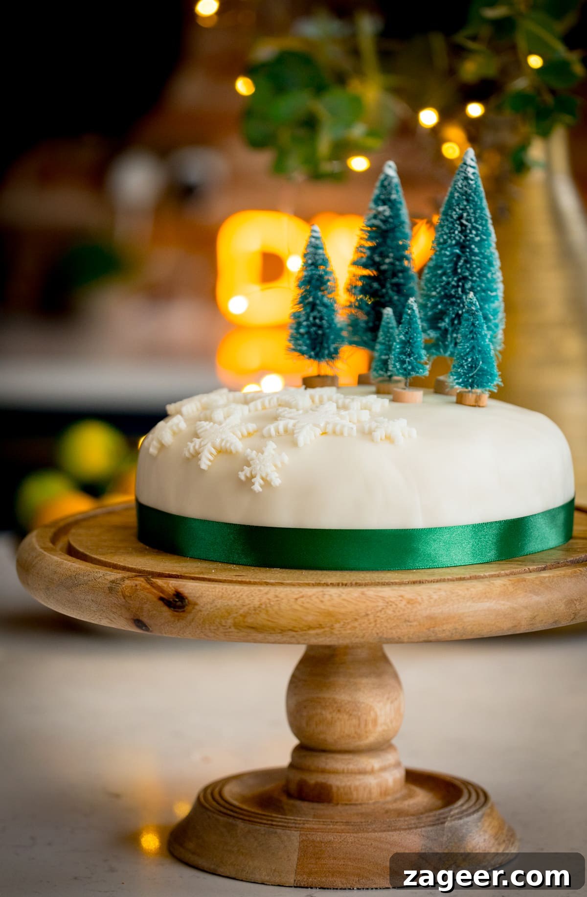 Christmas Cake Decorating Ideas for a Festive Finish 2 Tall side-on image showing a simple decorated Christmas cake - with a green ribbon surrounding the cake, white fondant snow flakes and mini Christmas trees on top. The cake is on a wooden cake stand on a kitchen surface with glowy lights and a bowl of fruit in the background.