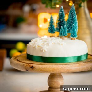 Christmas Cake Decorating Ideas for a Festive Finish 11 Square image showing a simple decorated Christmas cake - with a green ribbon surrounding the cake, white fondant snow flakes and mini Christmas trees on top. The cake is on a wooden cake stand on a kitchen surface with glowy lights and a bowl of fruit in the background.