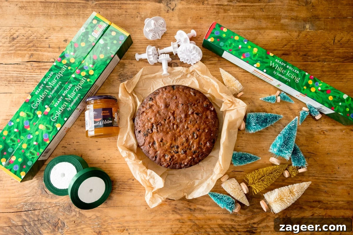 Christmas Cake Decorating Ideas for a Festive Finish 4 A fruitcake laying on baking parchment on a wooden table. The cake is surrounded by equipment and ingredients to decorate the cake as a Christmas cake. This includes rolls of marzipan, fondant icing, rolls of green ribbon, Apricot jam, snowflake fondant cutters and mini Christmas trees.