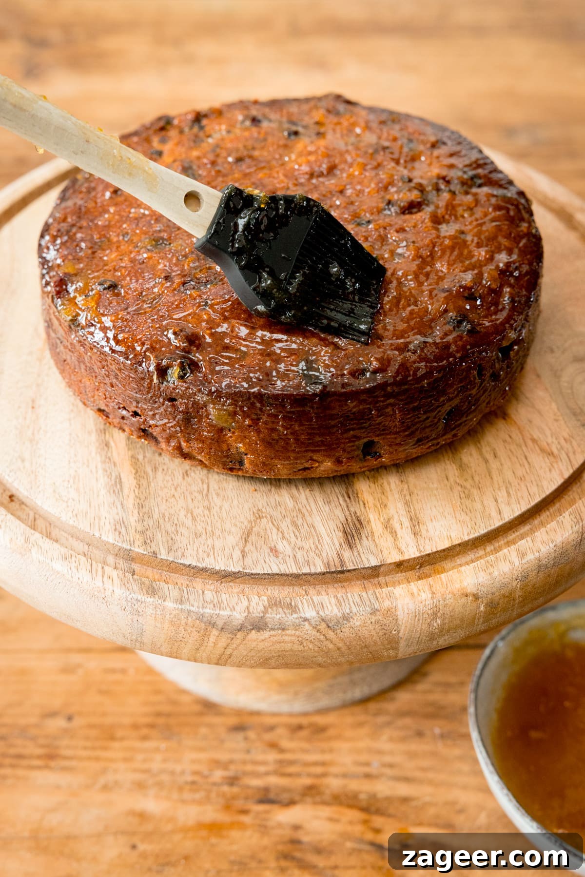 Christmas Cake Decorating Ideas for a Festive Finish 5 A fruitcake on a wooden cake stand, on a wooden table. The cake is being brushed with Apricot jam.