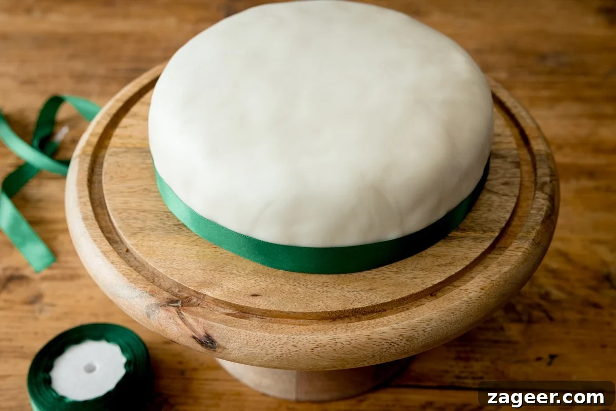 Christmas Cake Decorating Ideas for a Festive Finish 7 A fondant covered Christmas cake with a green ribbon on a wooden cake stand, on a wooden table. There is a roll or ribbon and a piece of ribbon next to the cake stand.