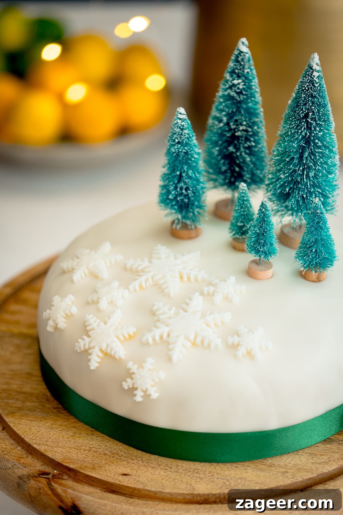 Christmas Cake Decorating Ideas for a Festive Finish 9 A close-up image of a simple decorated Christmas cake - with a green ribbon surrounding the cake, white fondant snow flakes and mini Christmas trees on top. The cake is on a wooden cake stand on a kitchen surface with glowy lights and a bowl of fruit in the background.