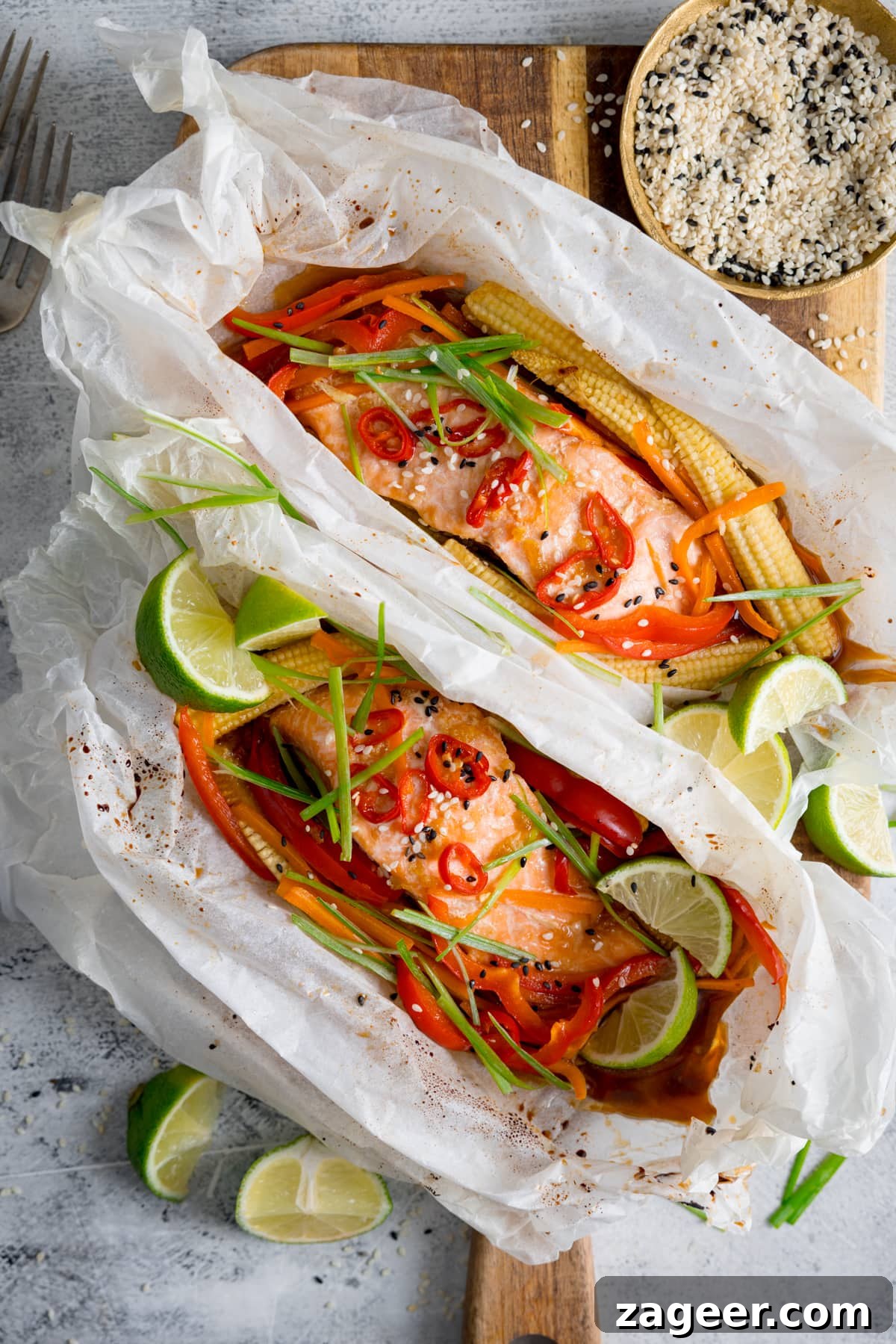 Salmon en Papillote: Steamed Baked Salmon Wrapped in Parchment 2 Overhead of two salmon fillets cooked in individual baking parchment parcels with slices of chilli, peppers, babycorn, carrot and spring onion. Also topped with wedges of lime. The parcels are open, on a wooden board on a light background. There is a dish of sesame seeds and some lime wedges nearby.