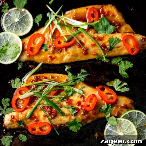 Salmon en Papillote: Steamed Baked Salmon Wrapped in Parchment 11 Two fillets of Asian Sea Bass, glazed and baked, topped with sliced red chilli, spring onions, and coriander, garnished with fresh lime slices on a dark background.