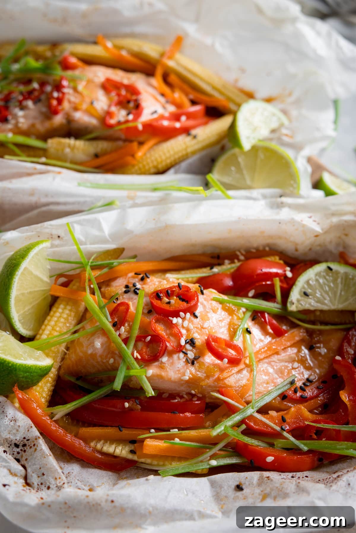 Salmon en Papillote: Steamed Baked Salmon Wrapped in Parchment 4 Close up overhead image of two salmon fillets cooked in individual baking parchment parcels with slices of chilli, peppers, babycorn, carrot and spring onion. Also topped with wedges of lime. The parcels are open.