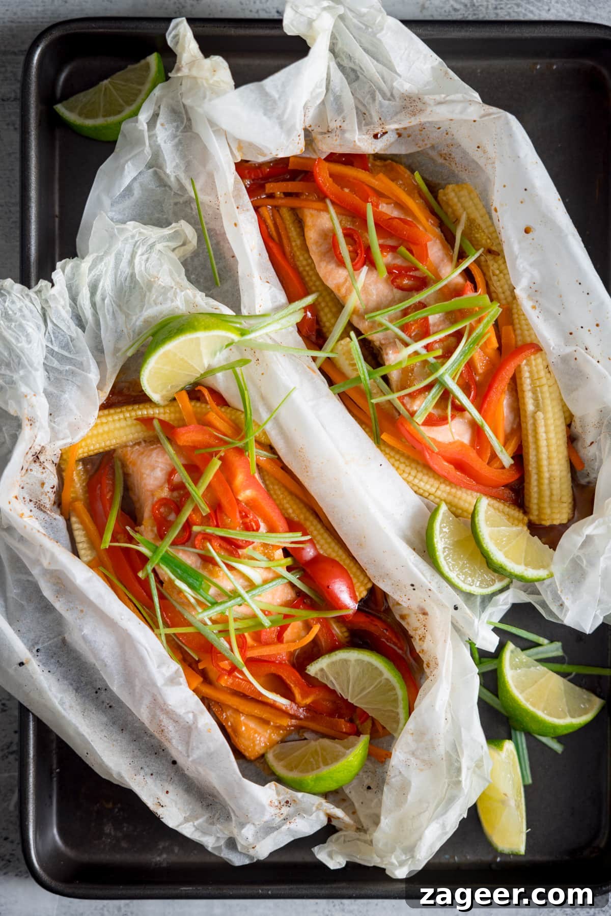 Salmon en Papillote: Steamed Baked Salmon Wrapped in Parchment 5 Two salmon fillets cooked in individual baking parchment parcels with slices of chilli, peppers, babycorn, carrot and spring onion. Also topped with wedges of lime. The parcels are open, on a dark baking tray. There are some lime wedges nearby.