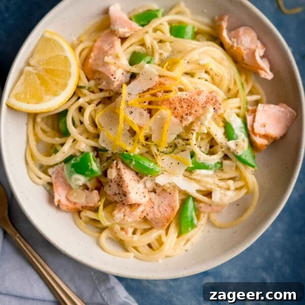 Salmon en Papillote: Steamed Baked Salmon Wrapped in Parchment 8 Square overhead image of salmon and spaghetti in a creamy lemon sauce in a white bowl with a lemon wedge in the bowl. The bowl is on a blue background. There is a fork and a napkin nearby.