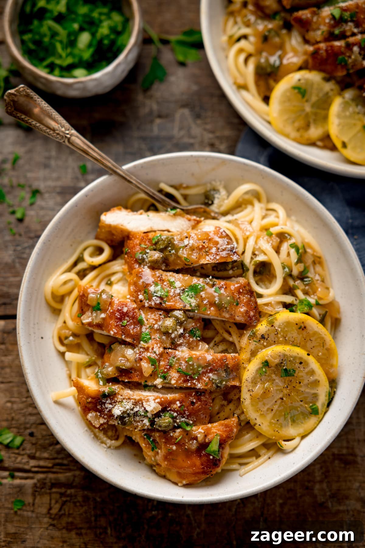 Tall overhead image of lemon chicken piccata on a bed of linguine with slices of lemon in a white bowl. The chicken has been sliced into strips and there is a fork sticking out. The bowl is on a wooden table next to a small bowl of herbs. There is a further bowl of lemon chicken piccata just out of shot at the top of the frame.