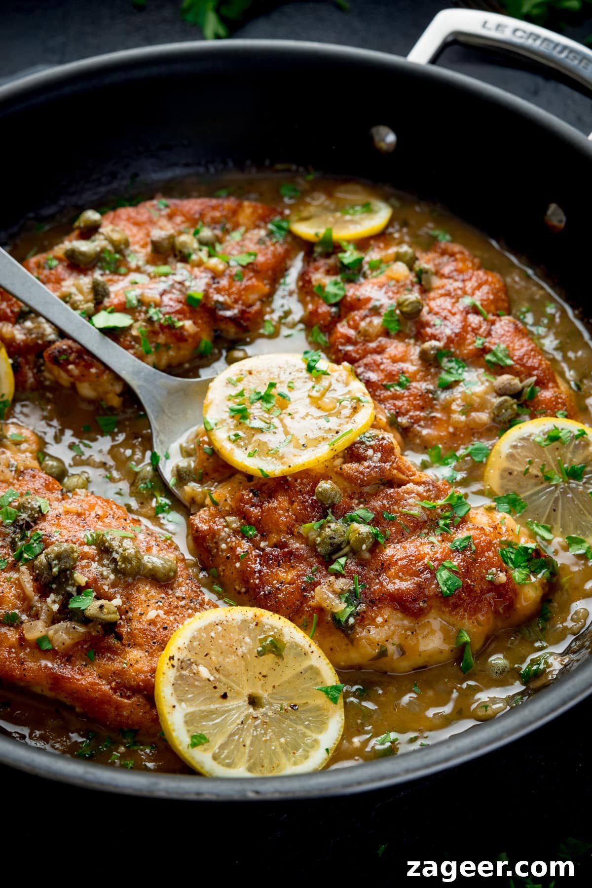 Lemon chicken piccata in a black pan topped with lemon slices and parsley. There is a spoon starting to lift up one of the pieces of chicken.