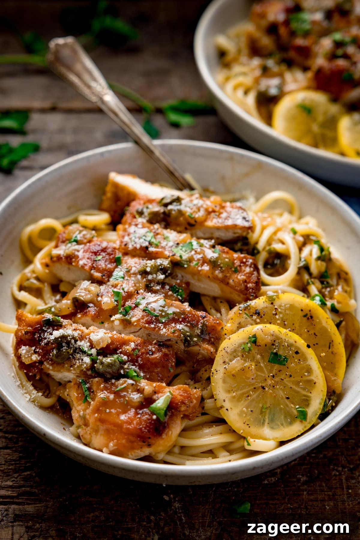 Tall image of lemon chicken piccata on a bed of linguine with slices of lemon in a white bowl. The chicken has been sliced into strips. The bowl is on a wooden table and there is a fork sticking out. There is a second bowl in the background.