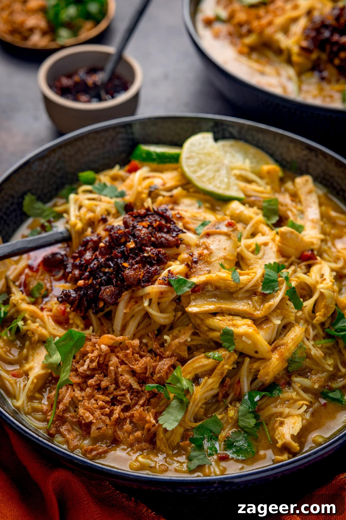 30-Minute Chicken Laksa Recipe: Flavorful Coconut Noodle Soup 2 Close up picture of some quick chicken laksa in a dark bowl, topped with crispy onions, chilli crisp and fresh coriander.