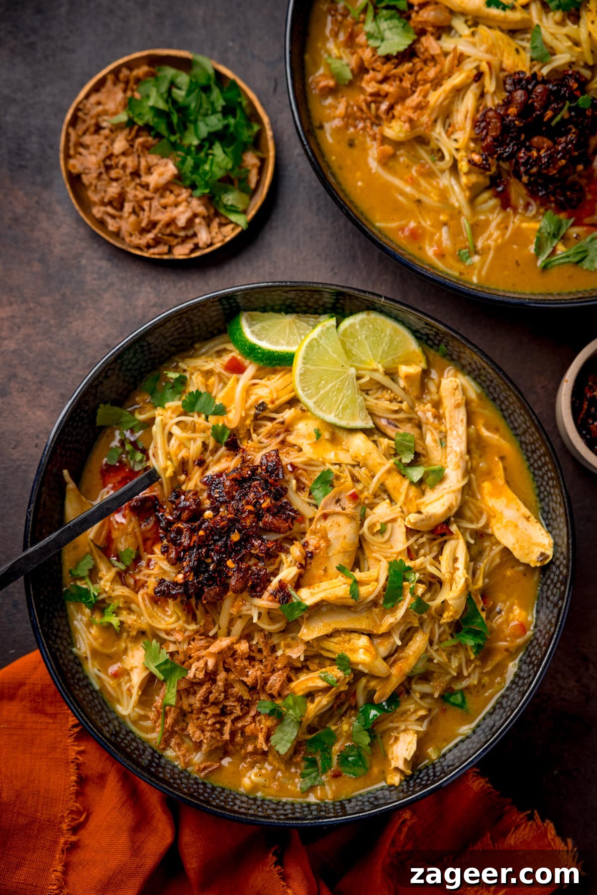 30-Minute Chicken Laksa Recipe: Flavorful Coconut Noodle Soup 5 Overhead picture of a black textured bowl full of chicken laksa topped with some chilli crisp and a sprinkling of fresh coriander and some lime wedges and an orange linen cloth in the foreground.