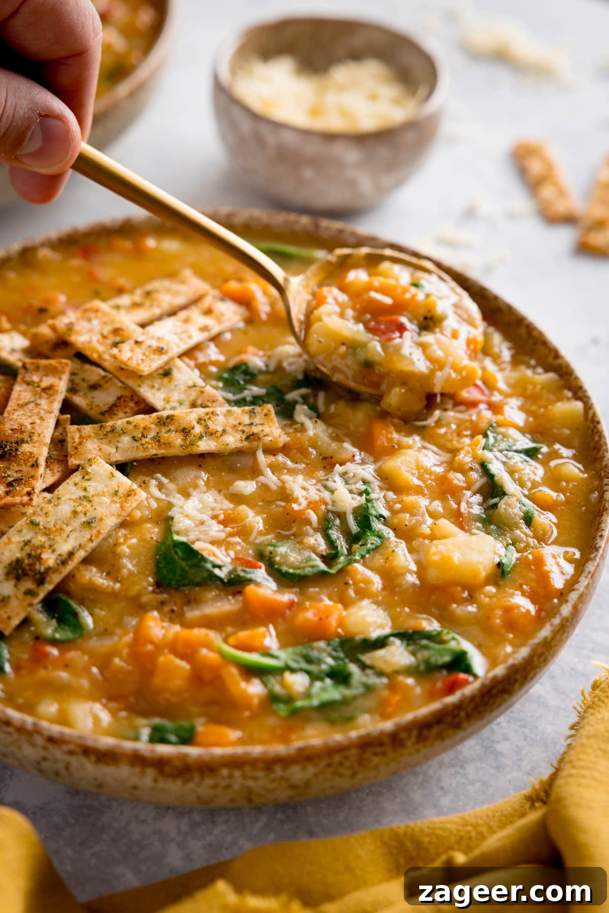 Hearty Slow Cooker Lentil Soup Recipe for Busy Weeknights 2 Tall image of red lentil soup topped with crispy tortilla strips in a brown speckled bowl. The bowl is on a light background. A spoonful of the soup is being lifted from the bowl. There are scattered tortilla strips and a small bowl of grated cheese at the top of the image and yellow napkin at the bottom of the image.