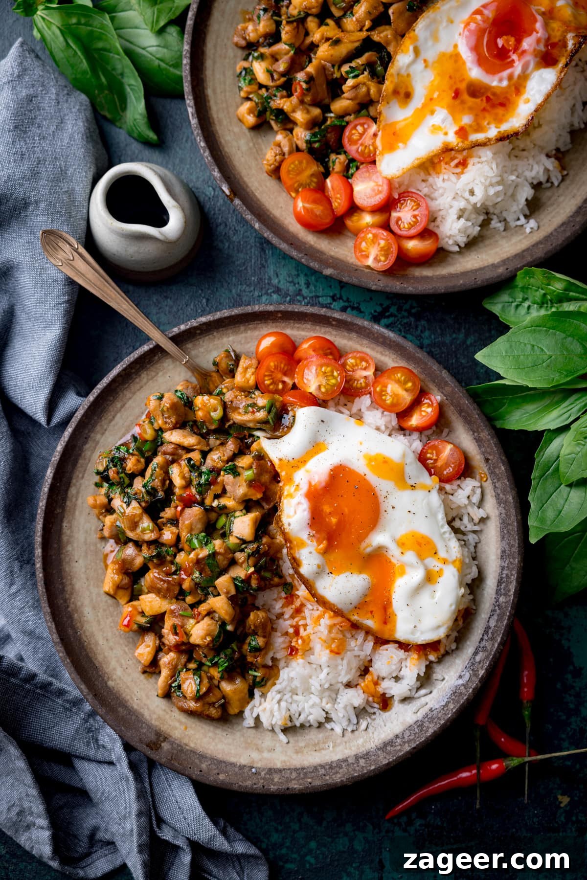 A tall overhead image of thai basil chicken served on a grey dish with rice that has a sunny side up egg on top and sliced cherry tomatoes on the top right of the dish. There is a copper fork sticking out of the dish. On the bottom right of the image is a bunch of thai basil. On the top right of the image is the corner of an identical bowl of thai basil chicken and a small white jug of sauce. In the background on the top left of the image is another bunch of basil and a grey handkerchief. there are a few red chillies scattered around. The image is set on a dark blue background.