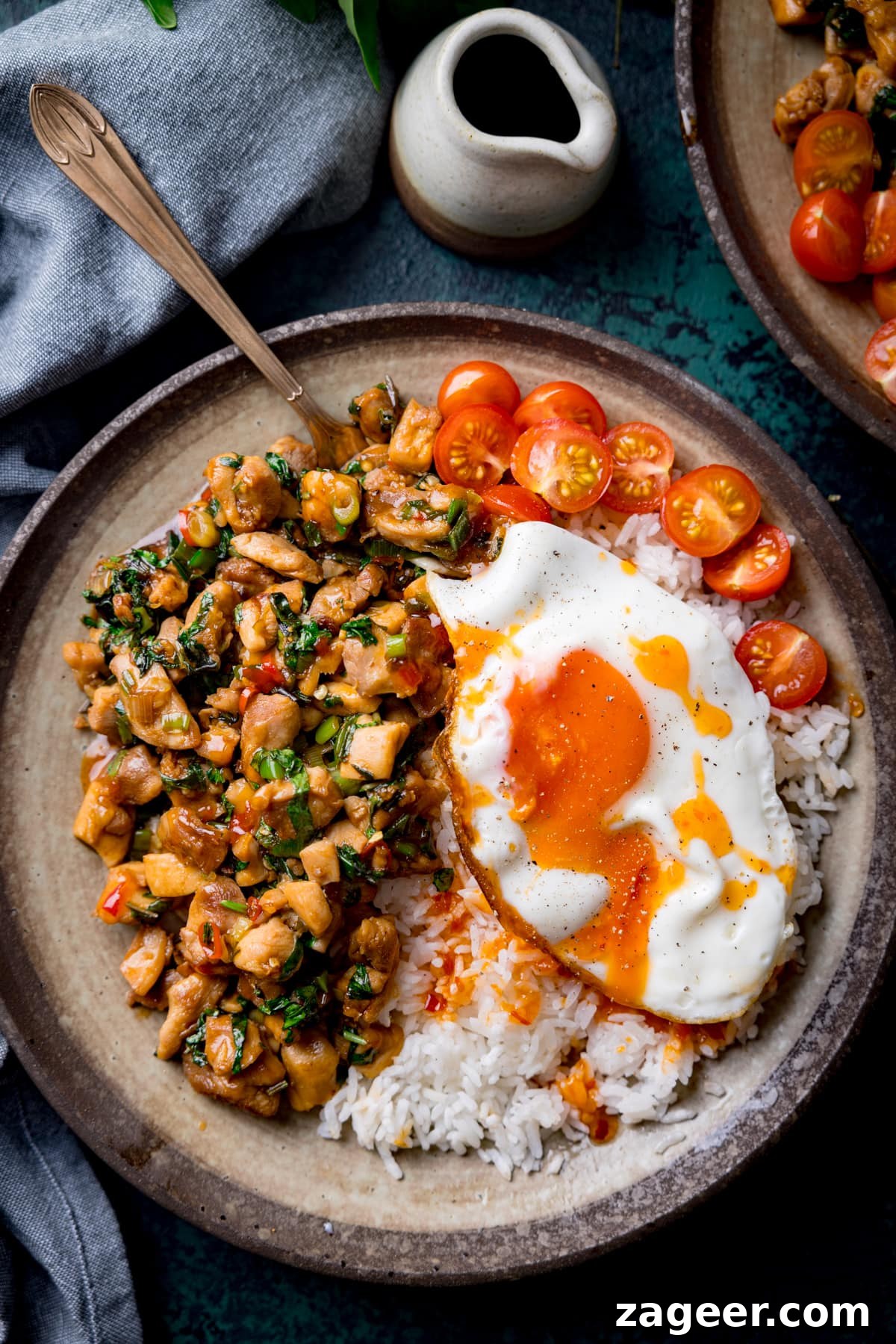 A tall overhead image of thai basil chicken served on a grey dish with rice that has a sunny side up egg on top and sliced cherry tomatoes on the top right of the dish. There is a copper fork sticking out of the dish. On the bottom right of the image is a bunch of thai basil. On the top right of the image is the corner of an identical bowl of thai basil chicken and a small white jug of sauce. In the background on the left of the image is a grey handkerchief. The image is set on a dark blue background.