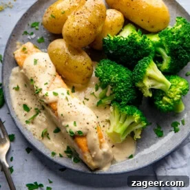 A plate with boiled potatoes, steamed broccoli, and pan fried salmon fillet topped with a creamy white sauce, garnished with chopped parsley.
