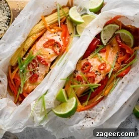 Square image of two salmon fillets cooked in individual baking parchment parcels with slices of chilli, peppers, babycorn, carrot and spring onion. Also topped with wedges of lime. The parcels are open, on a wooden board on a light background. There is a dish of sesame seeds nearby.