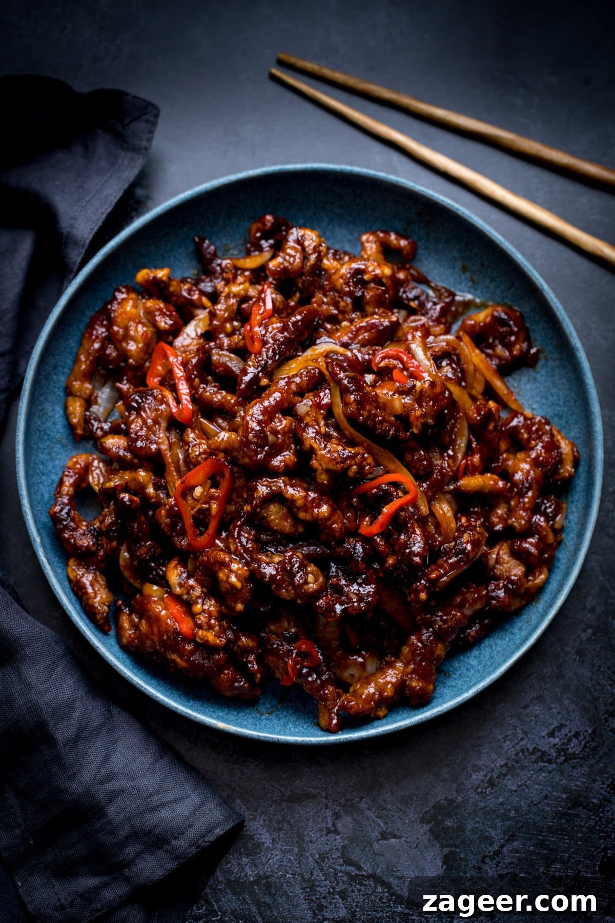 Crispy Chilli Beef Recipe with Sticky Sweet-Spicy Sauce 2 crispy chilli beef in a blue bowl on a dark background. Dark napkin and set of chopsticks also in shot.