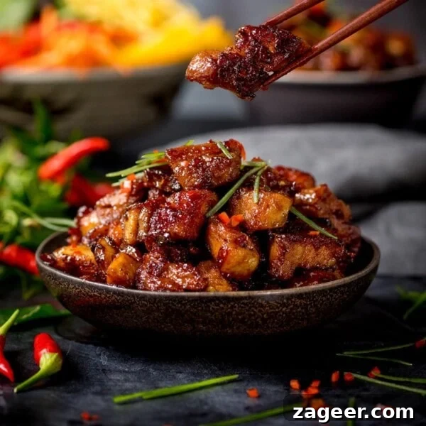 Crispy Chilli Beef Recipe with Sticky Sweet-Spicy Sauce 8 Close up image of bowl of Chinese sticky pork belly pieces. A piece is being taken from the bowl using a pair of chopsticks.