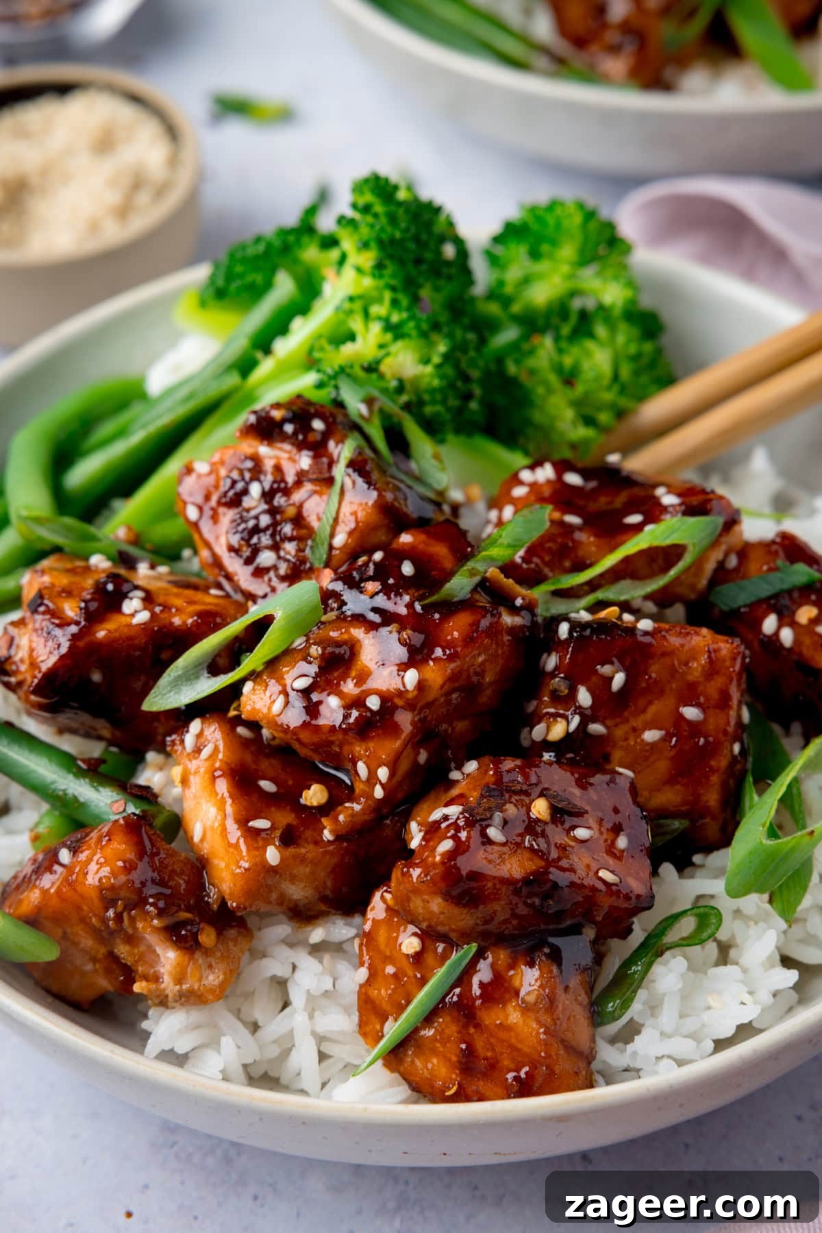 Crispy Air Fryer Teriyaki Salmon Recipe in 20 Minutes 2 A tall photo of Air Fryer Teriyaki Salmon served on white rice, with broccoli, and topped with sesame seeds and spring onions in a white bowl. There is a pair of wooden chopsticks sticking out of the dish to the right. In the top left of the background, there is a cream dish filled with white sesame seeds. On the top right of the background, you can see a white napkin and you can slightly see another white bowl of Air Fryer Teriyaki Salmon. This is all set on a white surface.
