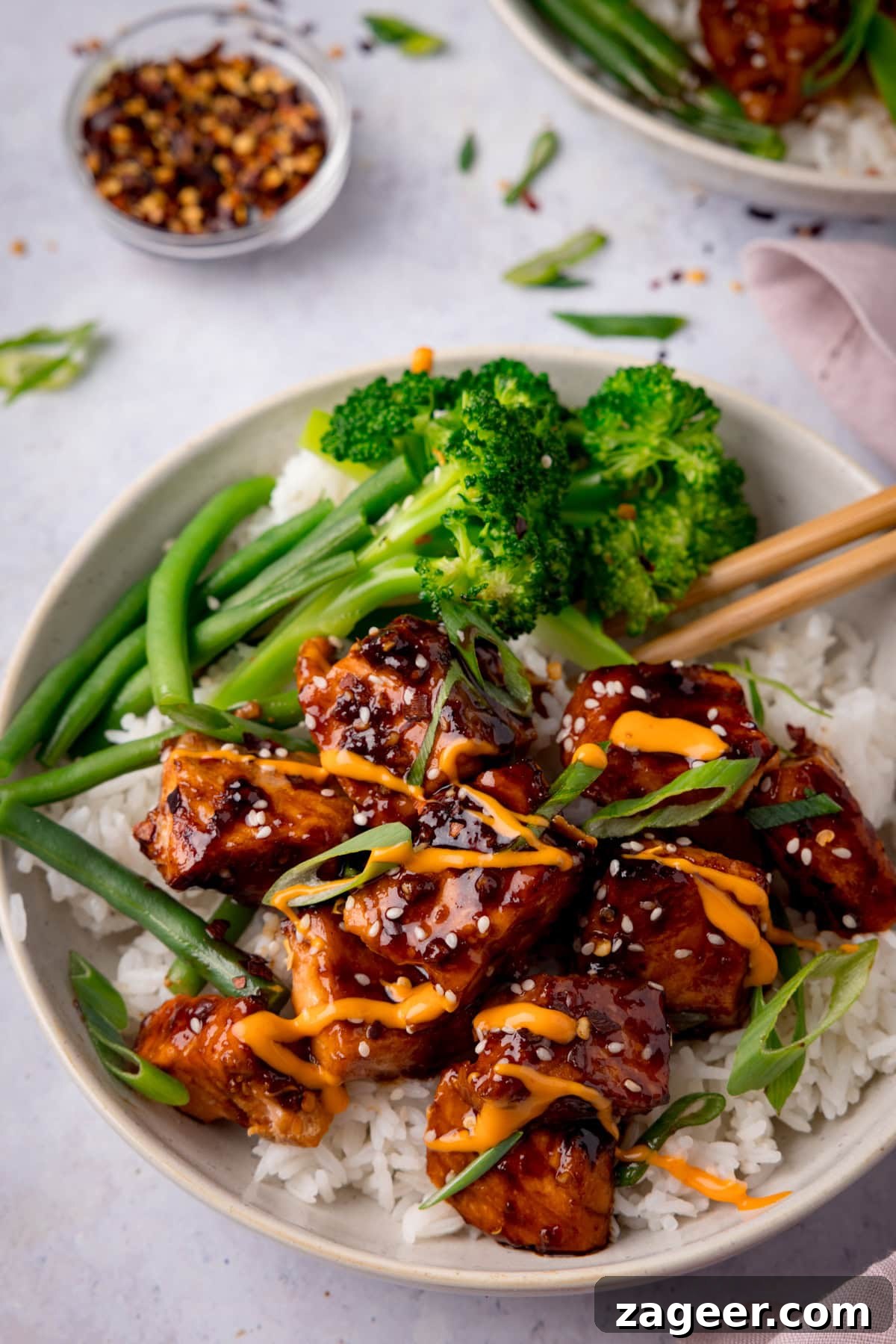 Crispy Air Fryer Teriyaki Salmon Recipe in 20 Minutes 4 A tall overhead photo of Air Fryer Teriyaki Salmon served on white rice, with broccoli, and topped with sesame seeds and spring onions in a white bowl. There is a pair of wooden chopsticks sticking out of the dish to the right. In the top left of the background, there is a white dish filled with dried chilli flakes. On the top right of the background, you can see a white napkin and you can slightly see another white bowl of Air Fryer Teriyaki Salmon. There are sesame seeds and spring onions scattered around. This is all set on a white surface.