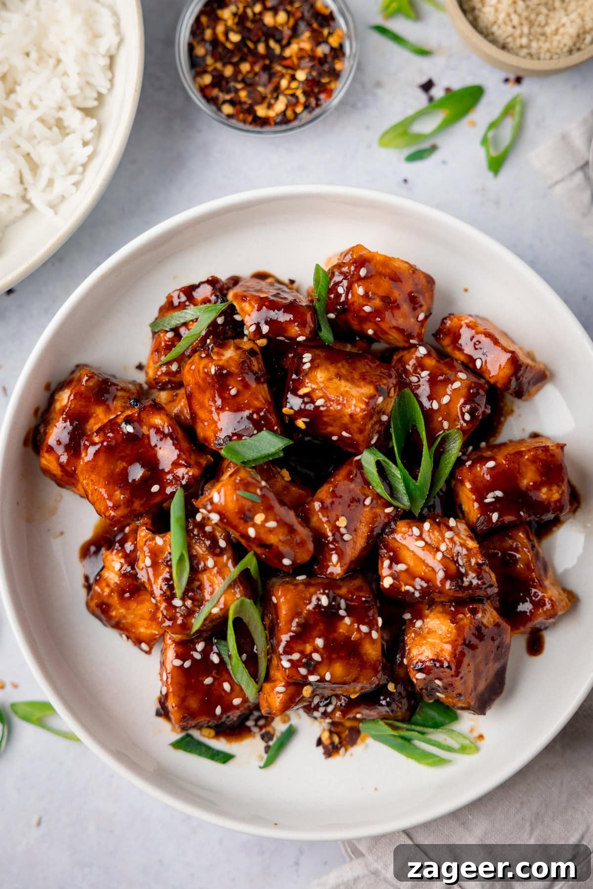 Crispy Air Fryer Teriyaki Salmon Recipe in 20 Minutes 6 A tall overhead shot of Air Fryer Teriyaki Salmon in a white bowl, with sesame seeds and spring onions on top and scattered around the surface. On the top left of the image you can see a white bowl of cooked white rice, and next to it a small clear dish of dried chilli flakes. And on the top right, there is a cream dish of sesame seeds. There is also a napkin beneath the main bowl of Air Fryer Teriyaki Salmon. This is set on a white background.