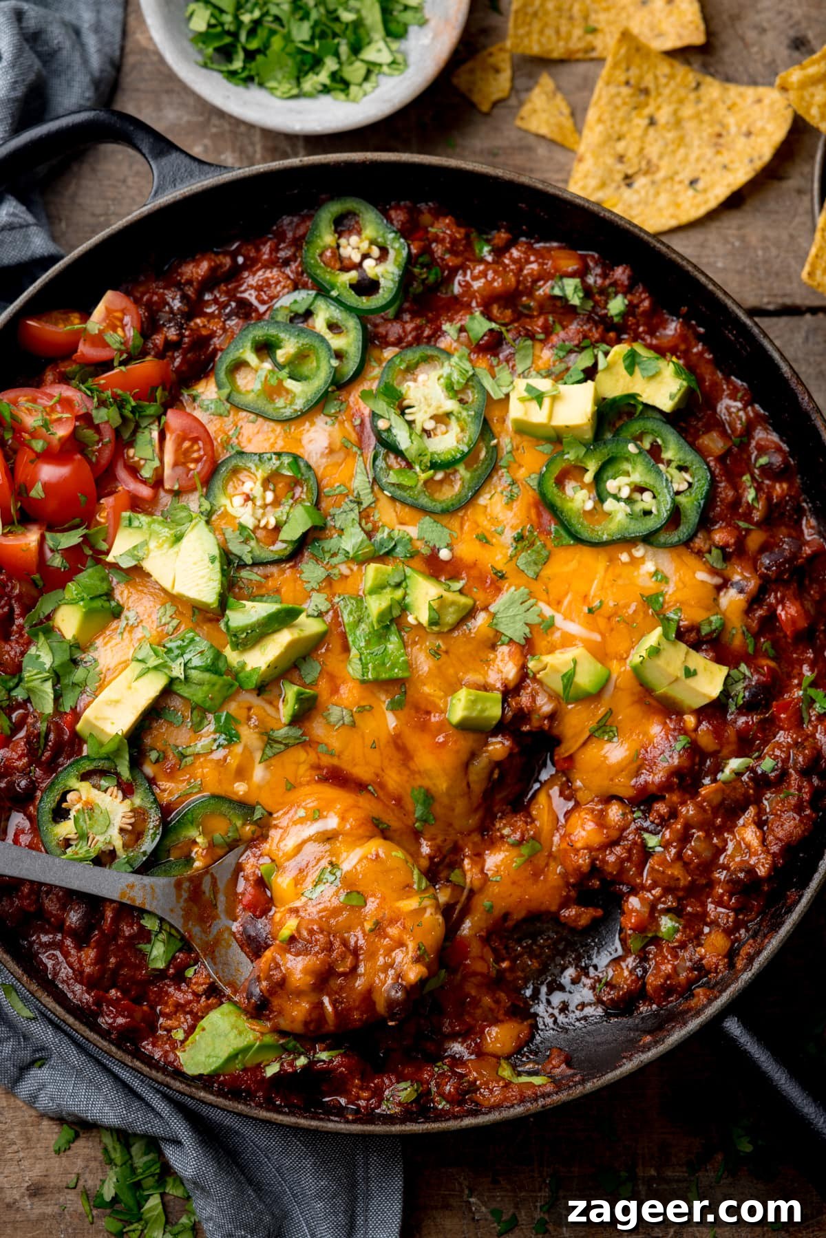 Tex‑Mex Minced Beef Recipe: Flavorful Beef for Tacos & Bowls 2 Tex-Mex minced beef in a black cast iron pan with cheese, jalapenos, coriander and avocados on top. There is a silver spoon sticking out of the dish. There is a grey dish of coriander on the top middle of the image, a grey napkin on the top left and some tortilla chips on the top right. This is on a wooden background.