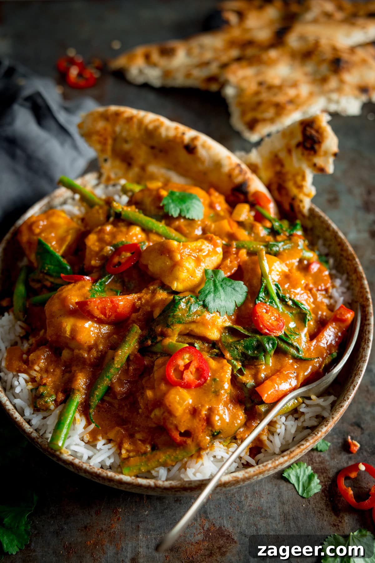 Creamy Monkfish Coconut Curry Recipe for Flavorful Dinner 2 Monkfish Coconut Curry on top of rice in a grey/brown bowl, topped with coriander and red chillies. There is a fork sticking out the bottom of the bowl and a couple of pieces of naan in the curry. This in on a grey background with a grey napkin. There is a piece of naan in the background as well as coriander scattered around.
