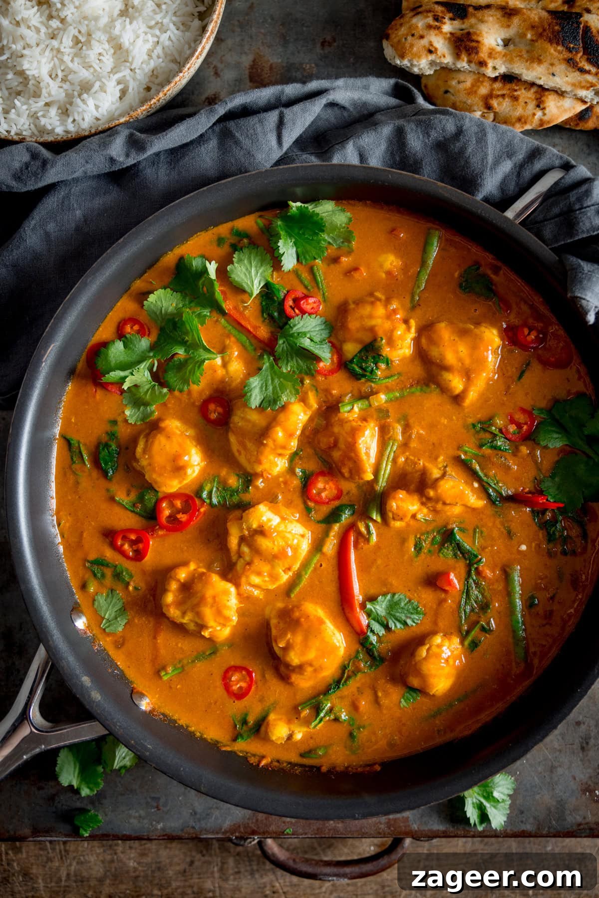 Creamy Monkfish Coconut Curry Recipe for Flavorful Dinner 5 A tall overhead shot of Monkfish Coconut Curry in a grey pan, topped with coriander. In the background on the top left there is a grey bowl filled with rice, and on the top righ there is 2 naan On a grey surface with a grey napkin in the background.