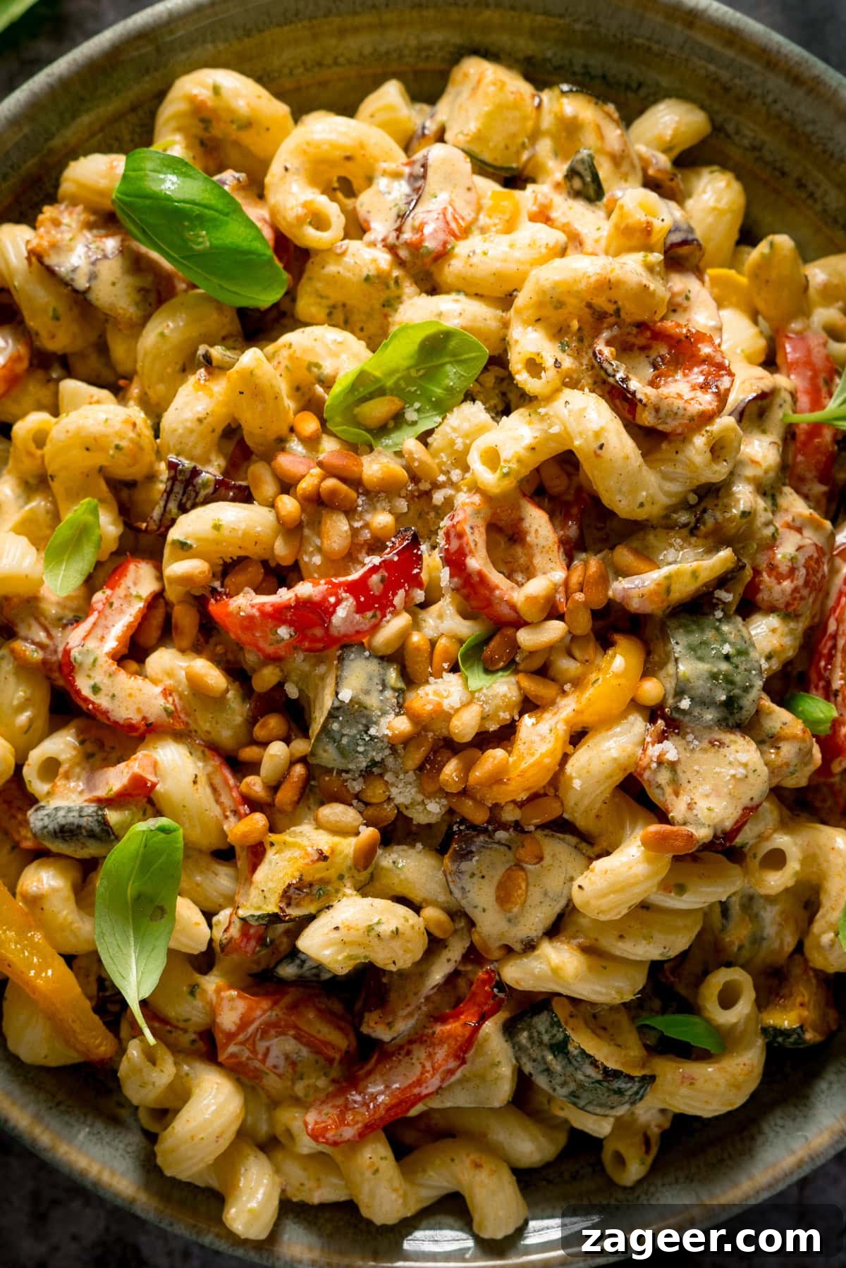 A very close-up tall image of roasted vegetable pesto pasta topped with toasted pine nuts, and a few small basil leaves. This is in a light grey bowl. The pasta an the bowl fill the whole image, so nothing else can be seen.