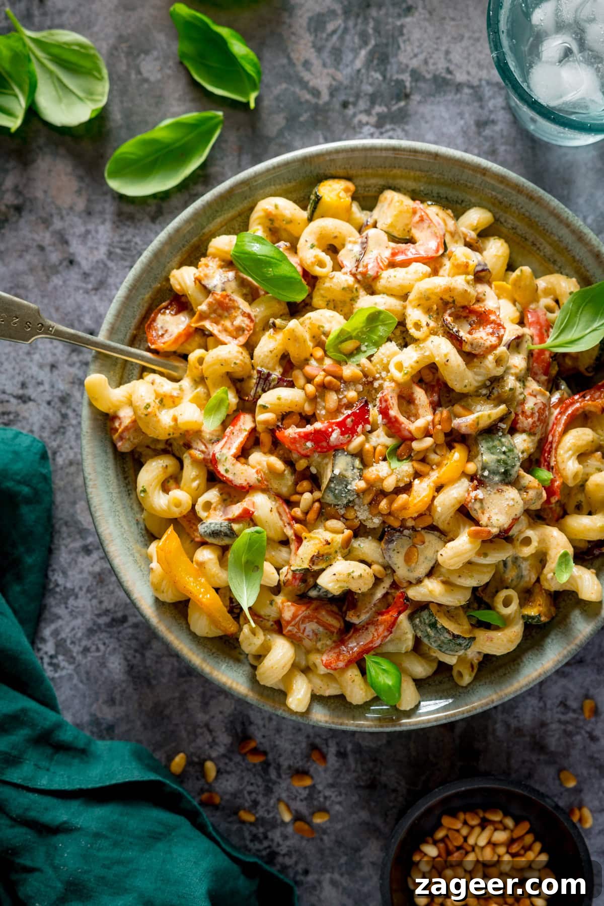 A tall, overhead image of roasted vegetable pesto pasta, topped with small basil leaves, and some toasted pine nuts, in a light blue/grey bowl, with a silver fork sticking out of the pasta. On the top right of the image, there is a glass of water with ice, on the top left there are a few medium-sized basil leaves scattered around. On the bottom right there is a small black dish of toasted pine nuts, with more toasted pine nuts scattered around. On the bottom left, there is a teal/green napkin. This is all placed on a grey background.