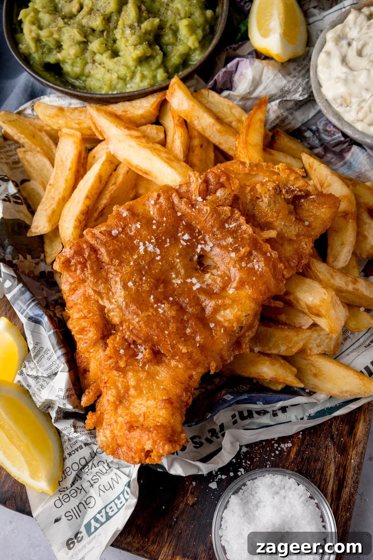Crispy Beer-Battered Fish Recipe for Perfect Fried Fish 2 A tall, overhead shot of beer-battered fish. There is one beer-battered fish fillet, topped with flakes of salt, on a bed of chips. The food is on top of a newspaper. To the left of the fish and chips are two wedges of lemon. In the top left of the image, there is a black bowl of mushy peas, with another wedge of lemon to the right of it. In the top right of the image, there is a grey dish of tartare sauce. This is all on a wooden board. On the bottom right of the wooden board, there is a clear dish of sea salt. This is all placed on a white marble background.