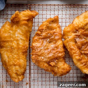 Crispy Beer-Battered Fish Recipe for Perfect Fried Fish 14 A square, overhead shot of three fish fillets, battered in the beer batter. There are three golden battered fish fillets laid side by side on a copper wire rack. They are topped with flakes of salt. The wire rack is on a white marble background, and in the top left corner, you can slightly see a blue napkin.