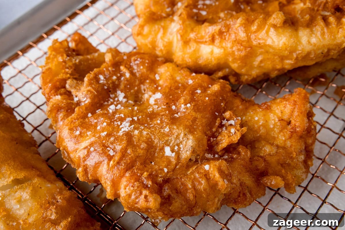 Crispy Beer-Battered Fish Recipe for Perfect Fried Fish 5 A wide, close-up shot of three fish fillets, battered in the beer batter. There are three golden battered fish fillets laid side by side on a copper wire rack. They are topped with flakes of salt. The wire rack is on a white marble background.